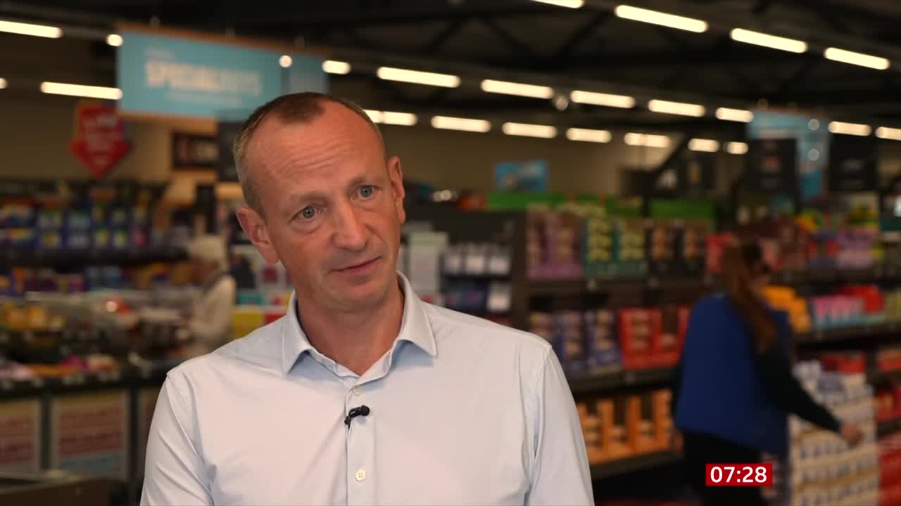 A man in a light blue shirt speaks into a microphone, likely being interviewed on BBC News. Behind him, shoppers browse the shelves of a supermarket in the United Kingdom.
