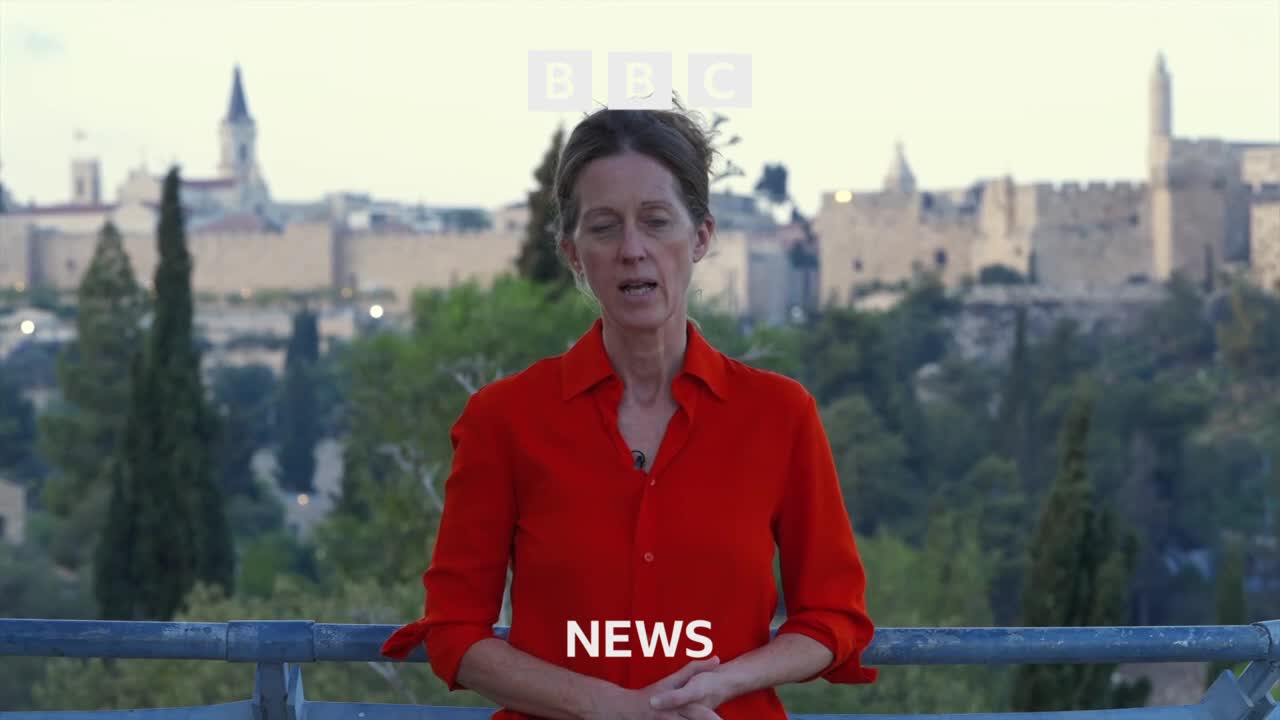 A woman in a red shirt stands before a historic cityscape, speaking directly to the camera. The BBC logo is visible above her head, and the word "NEWS" appears below.

