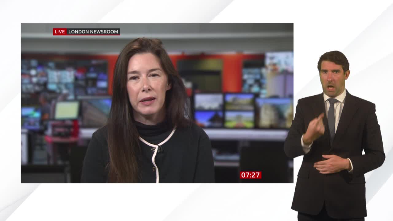 A woman with long brown hair speaks on a BBC News broadcast from the London newsroom. Beside her, a man in a suit interprets the news in sign language.
