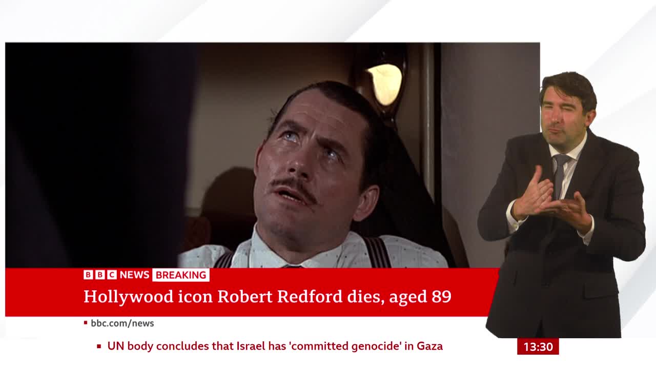 A BBC News graphic announces the death of Robert Redford, while a man in a suit signs in front of a screen. The image of the actor, with a mustache, is from a film.
