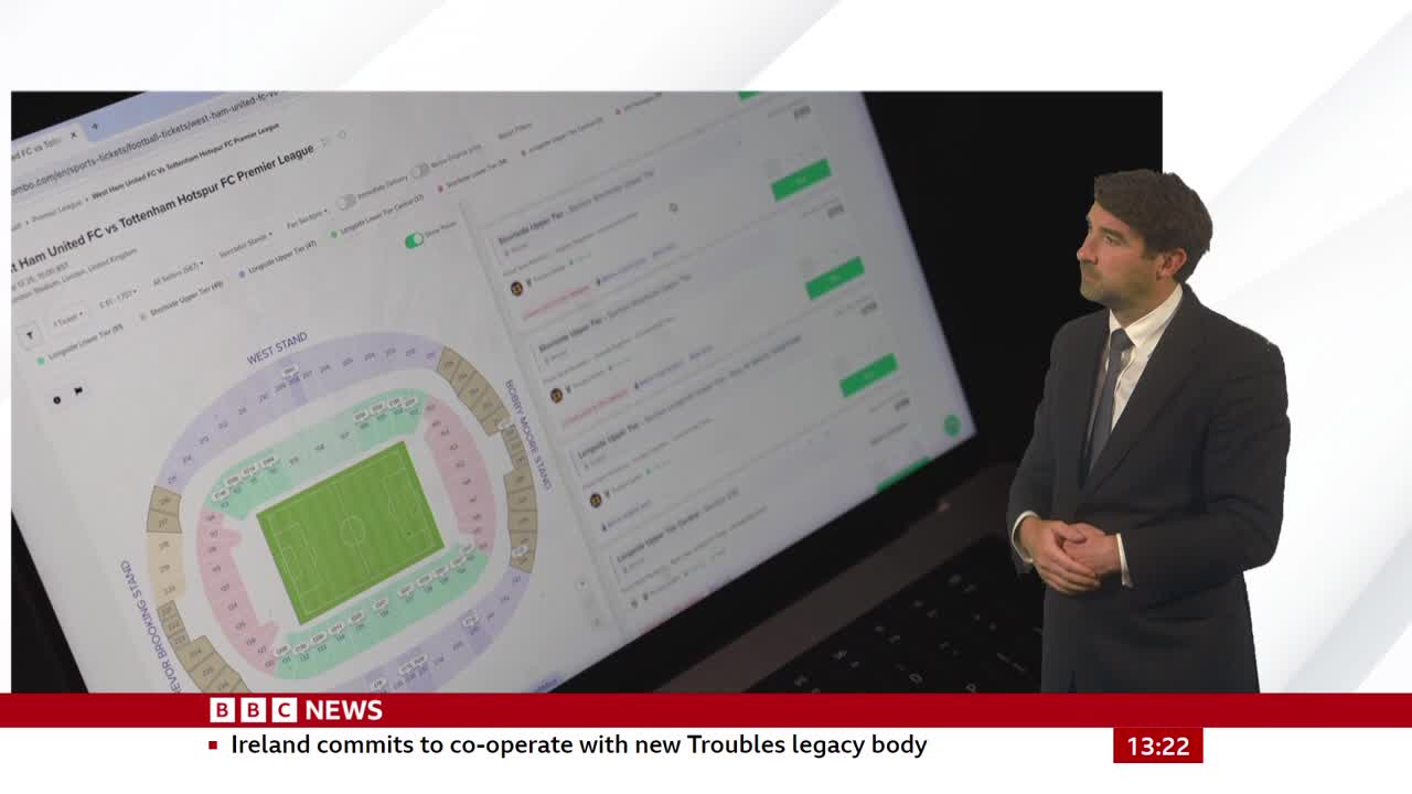 A laptop screen displays a stadium seating chart, with a man in a suit standing beside it. The BBC News logo and a headline about Ireland are visible at the bottom of the screen.

