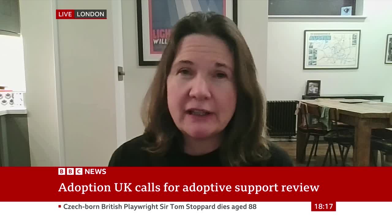 A woman in a black top is speaking, framed by a BBC News graphic about adoption support. Behind her, a dining table and framed artwork are visible in what appears to be a home in the United Kingdom.
