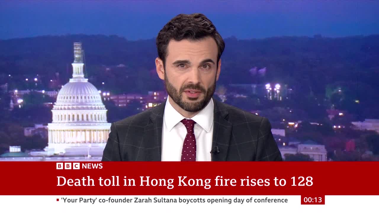 A BBC News anchor, wearing a suit and tie, is speaking directly to the camera. Behind him, a nighttime cityscape is visible, with the dome of a building illuminated in the background.
