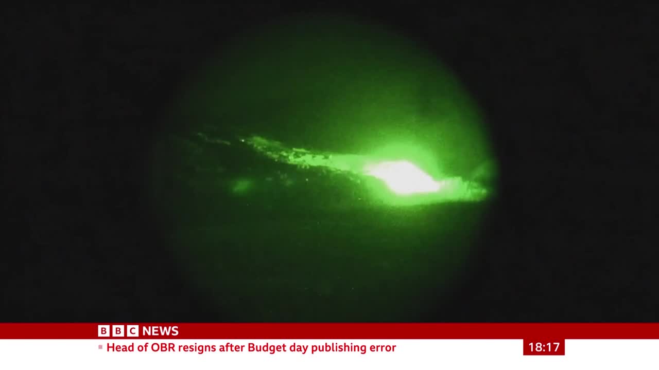 A bright green light illuminates a dark space, with a jagged, luminous streak across the center. The BBC News logo and a headline about a resignation are visible at the bottom of the screen.
