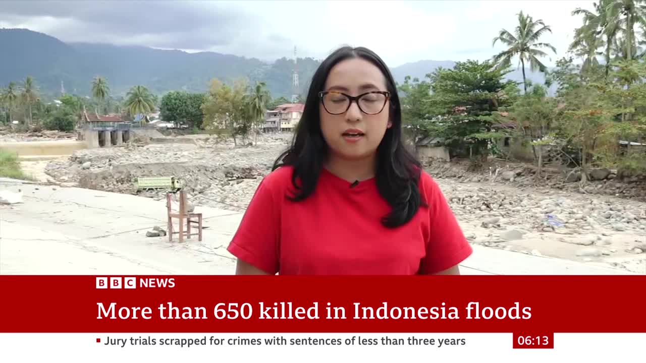 A BBC News correspondent stands on a concrete surface, speaking to the camera against a backdrop of devastation. Behind her, the remnants of a flooded area are visible, with debris and damaged structures.
