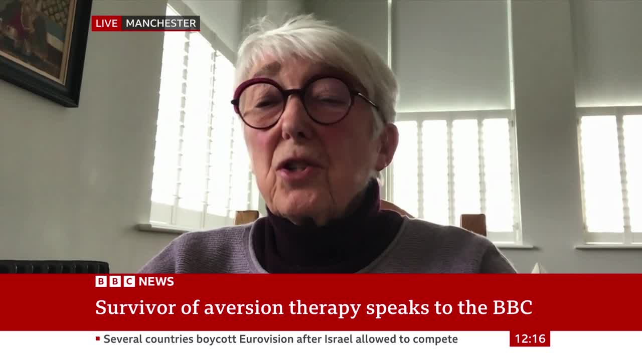 A woman in glasses is speaking, her mouth moving as she addresses the camera. The BBC News graphic identifies her as a survivor of aversion therapy, live from Manchester.
