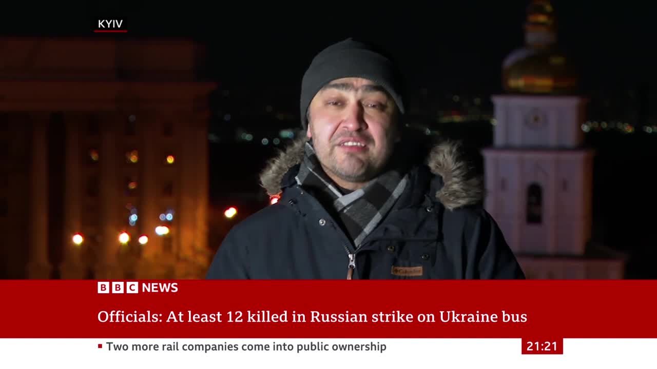 A man stands in front of a building at night, speaking to the camera. A BBC News chyron states that at least 12 people were killed in a Russian strike on a Ukrainian bus.