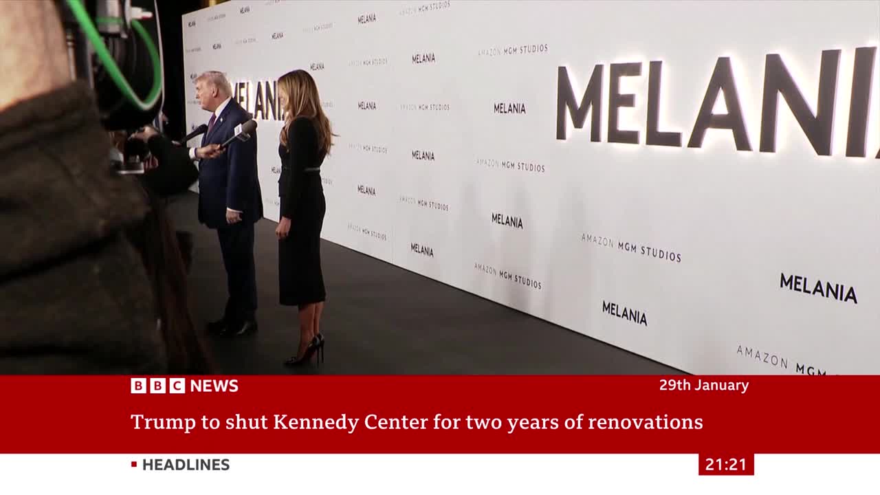 A man in a suit and a woman in a black dress stand on a red carpet, facing a wall with large "MELANIA" lettering. A camera operator is positioned to the left, filming the scene.