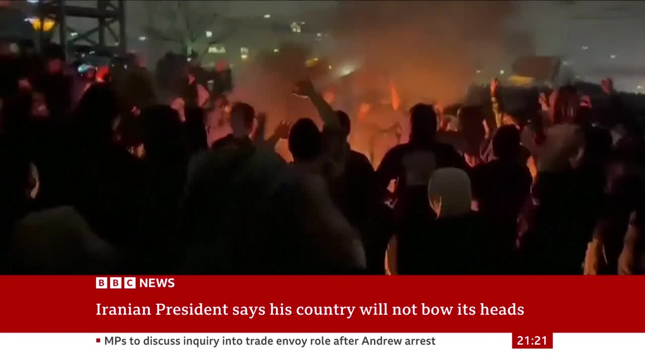 A crowd surges, silhouetted against a bonfire's glow. The BBC News ticker reports on the Iranian President's defiant stance. A crowd surges, silhouetted against a bonfire's glow. The BBC News ticker reports on the Iranian President's defiant stance.