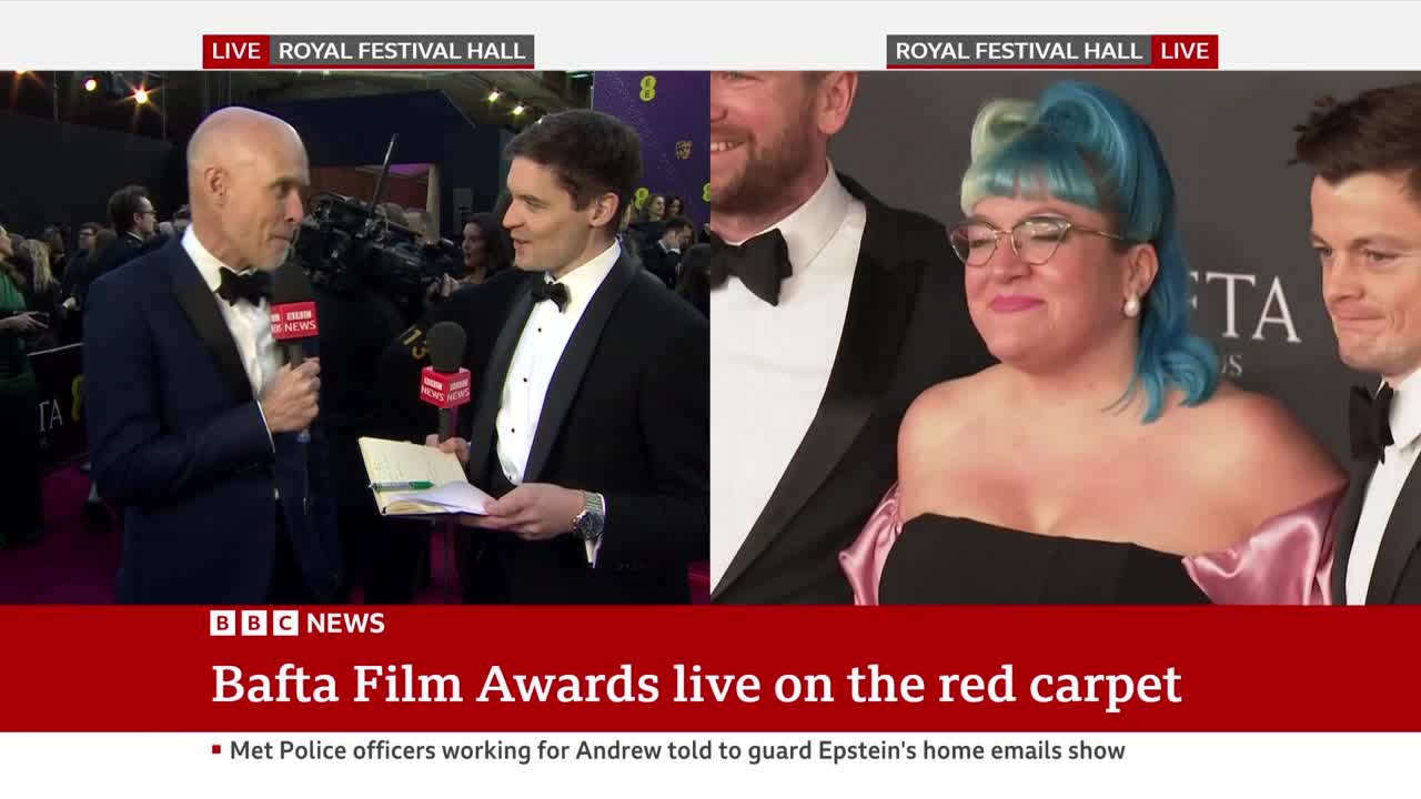 A BBC News reporter interviews a man in a tuxedo on a red carpet at the BAFTA Film Awards. A woman with bright blue hair and a man in a tuxedo stand nearby. A BBC News reporter interviews a man in a tuxedo on a red carpet at the BAFTA Film Awards. A woman with bright blue hair and a man in a tuxedo stand nearby.
