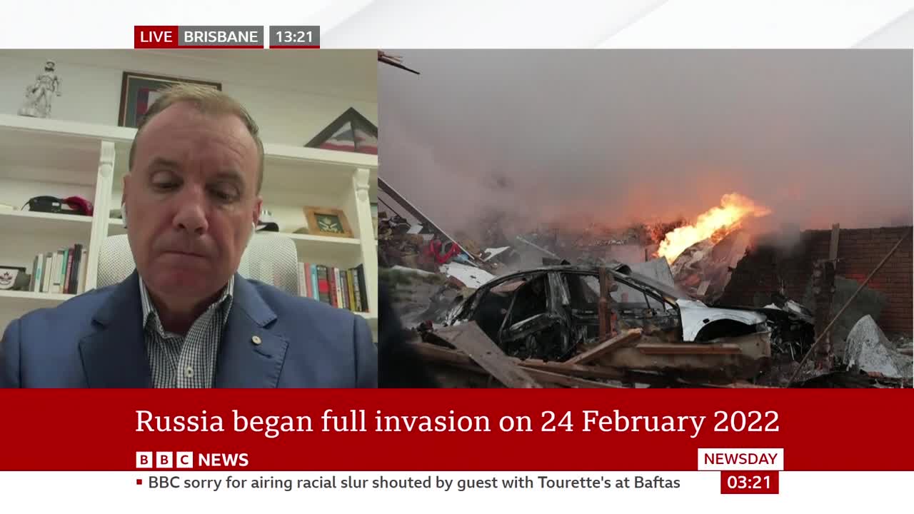 Flames erupt from a destroyed building, with a mangled car in the foreground. A man in a suit speaks on a screen, his expression serious. Flames erupt from a destroyed building, with a mangled car in the foreground. A man in a suit speaks on a screen, his expression serious.