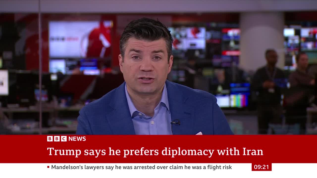 A news anchor in a blue suit speaks directly to the camera, with multiple screens displaying graphics behind him. A red banner at the bottom of the screen reads "Trump says he prefers diplomacy with Iran" and the BBC News logo. A news anchor in a blue suit speaks directly to the camera, with multiple screens displaying graphics behind him. A red banner at the bottom of the screen reads "Trump says he prefers diplomacy with Iran" and the BBC News logo.