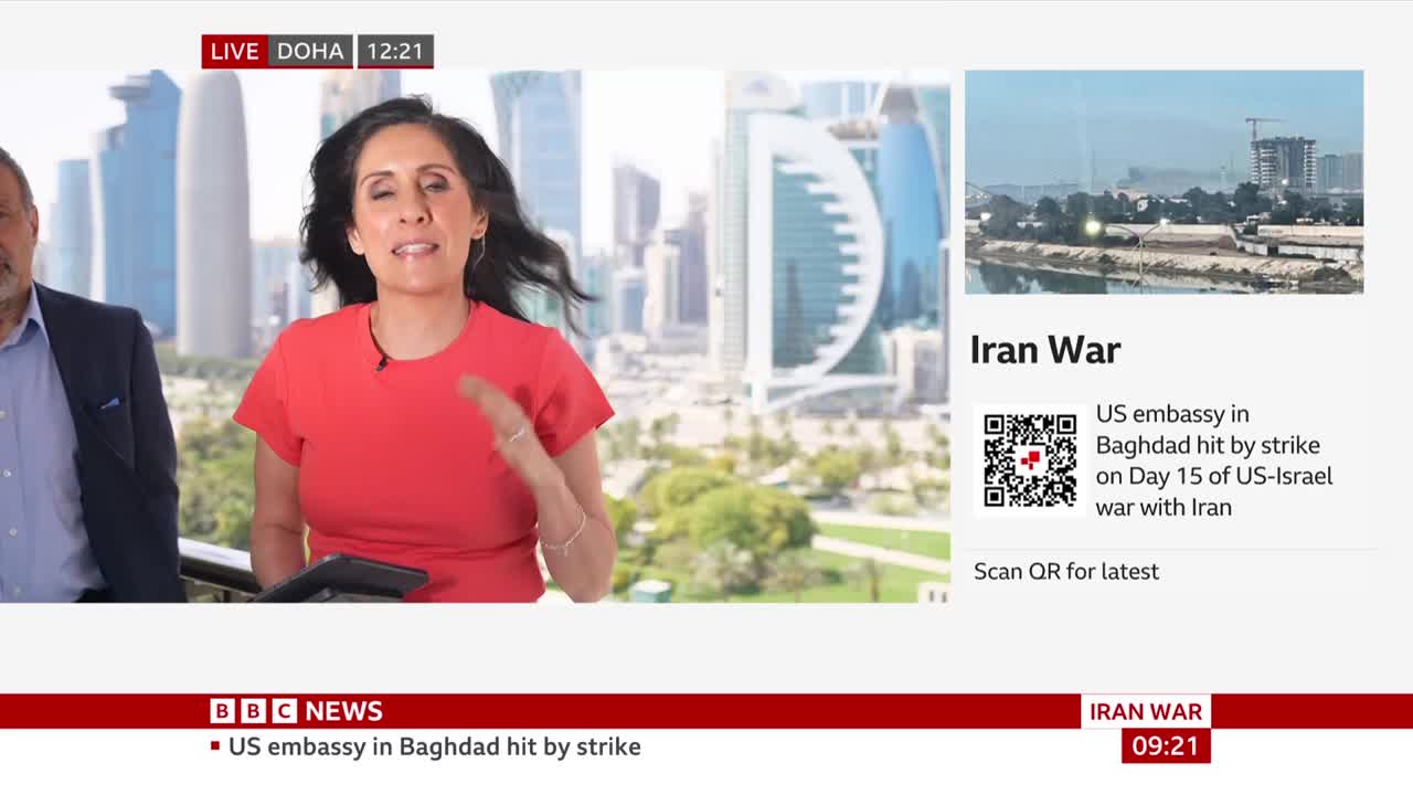 A woman in a red shirt speaks animatedly, gesturing with her right hand. Behind her, a cityscape with modern skyscrapers stretches under a bright sky. The BBC News chyron at the bottom indicates a report on the "Iran War."