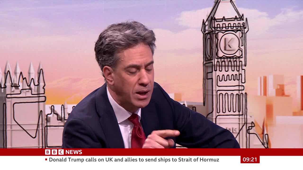 A man in a suit and tie gestures with his right hand as he speaks. Behind him, a stylized line drawing of Big Ben and the Houses of Parliament fills the screen.