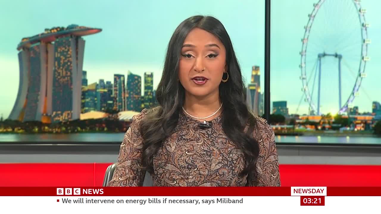 A BBC News presenter speaks directly to the camera, with a cityscape featuring a large Ferris wheel behind her. The ticker at the bottom of the screen reads "We will intervene on energy bills if necessary, says Miliband."