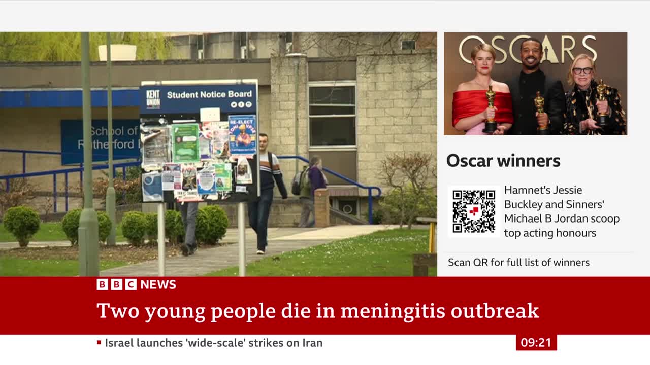 A young man walks past a busy student notice board outside a building. Another person heads up some steps in the background.