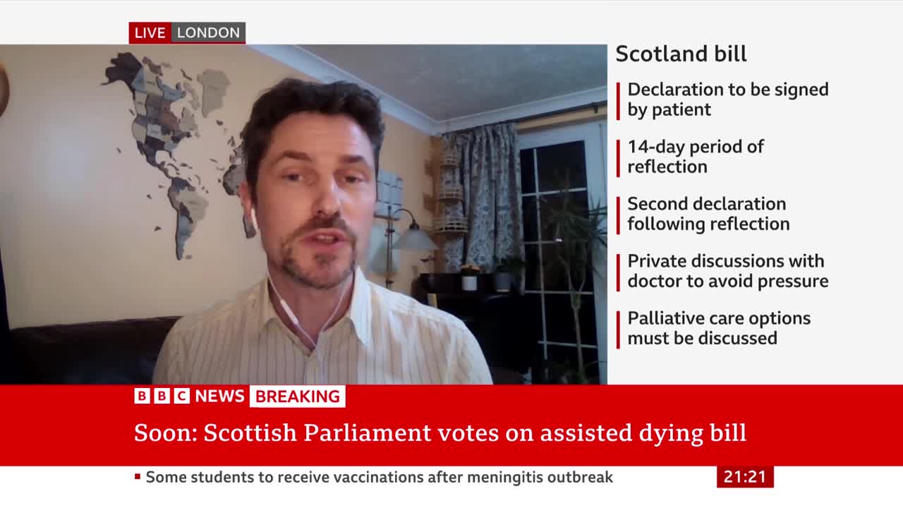 A man speaks directly to the camera, a world map on the wall behind him. A BBC News banner announces a vote on an assisted dying bill in the Scottish Parliament.