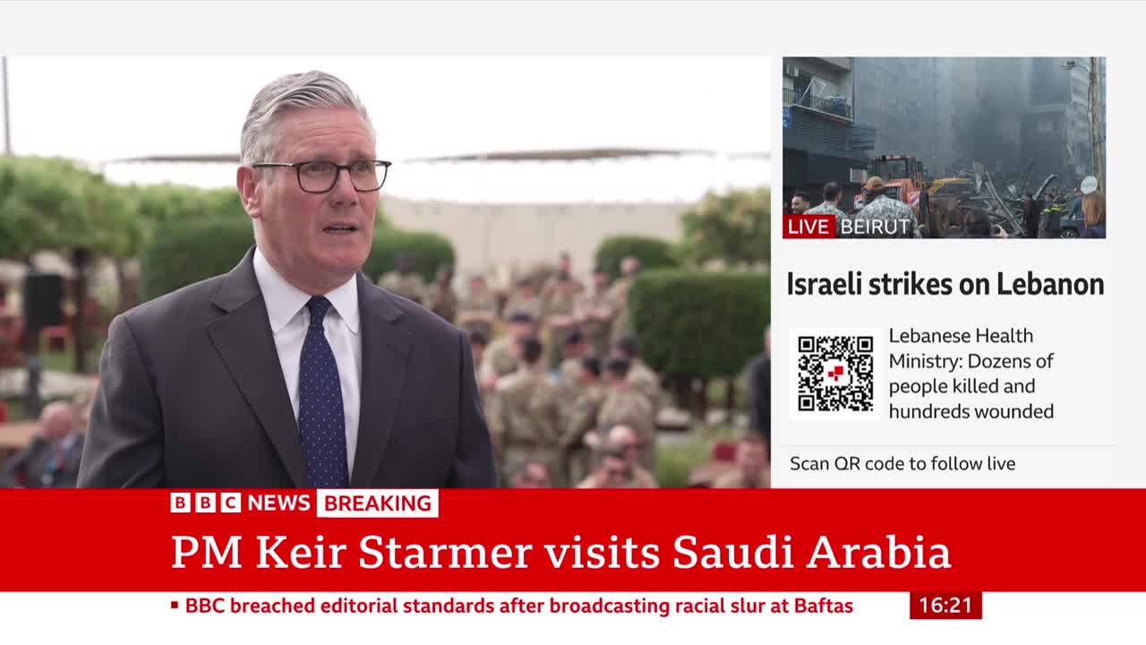 Keir Starmer, in a dark suit and tie, speaks to the camera. Behind him, a group of soldiers stand at attention. A BBC News banner at the bottom of the screen announces "PM Keir Starmer visits Saudi Arabia."
