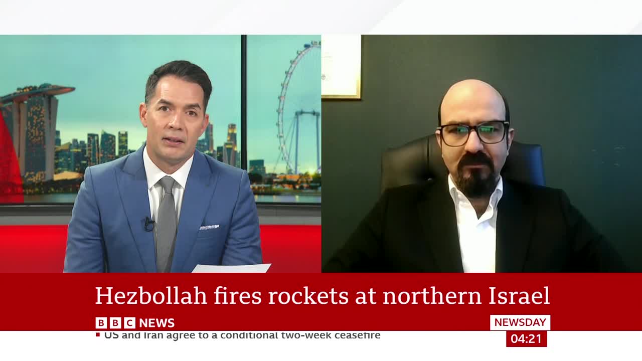 A news anchor in a blue suit looks directly at the camera, while a man in a suit and glasses appears on a split screen. A red banner across the bottom of the screen reads "Hezbollah fires rockets at northern Israel."