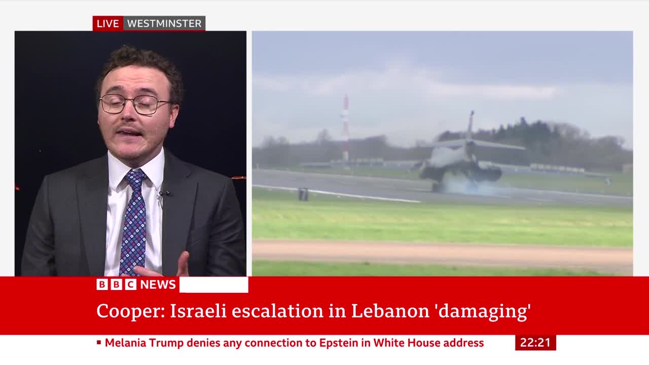 A military jet touches down on a runway, kicking up a cloud of dust. The BBC News broadcast shows this alongside a commentator speaking live from Westminster.