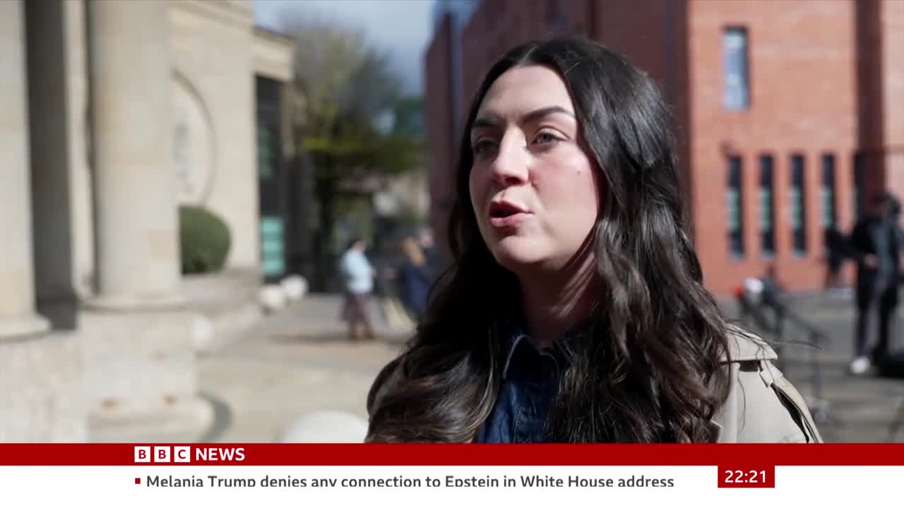 A woman speaks directly to the camera, her voice clear as she discusses news from the UK. Behind her, people move past on a sunny day, their figures slightly blurred against the backdrop of brick buildings.