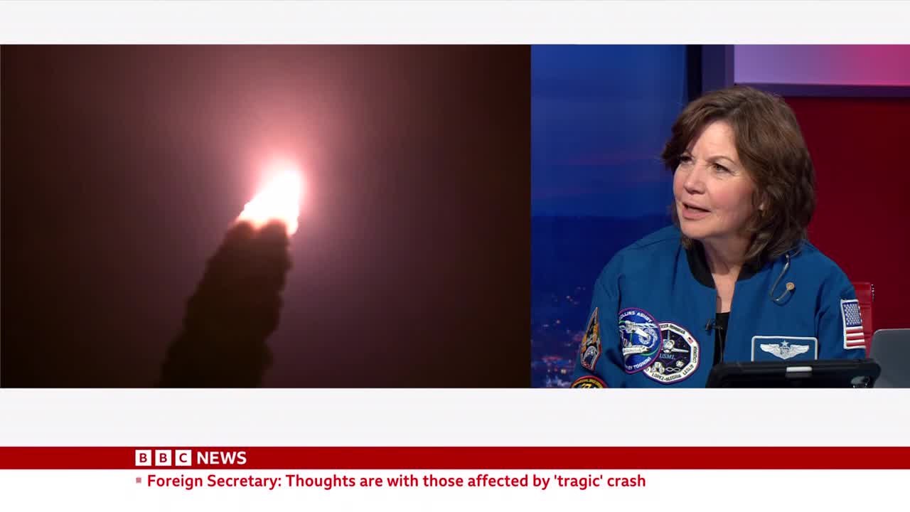 A rocket ascends into the sky, trailing a thick plume of smoke illuminated by its bright engine. On a screen beside it, a woman in a blue flight suit speaks, her expression serious.