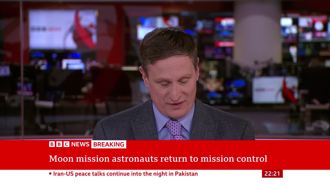 A man in a suit and tie looks down, speaking. Behind him, screens display news graphics, including a BBC News banner announcing "Moon mission astronauts return to mission control."
