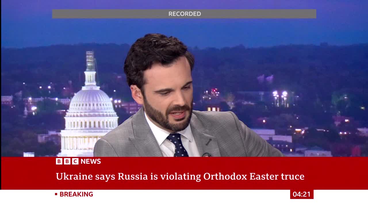 A man in a suit speaks, his gaze directed slightly off-camera. Behind him, the dome of the US Capitol building is illuminated against a twilight sky.