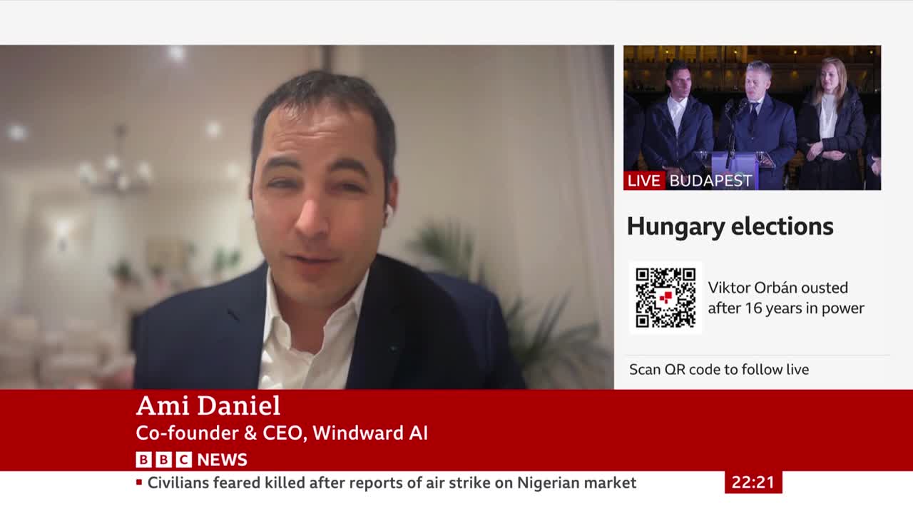 A man in a suit and white shirt is speaking, with a plant behind him. To the right, a live feed from Budapest shows three people at a podium.