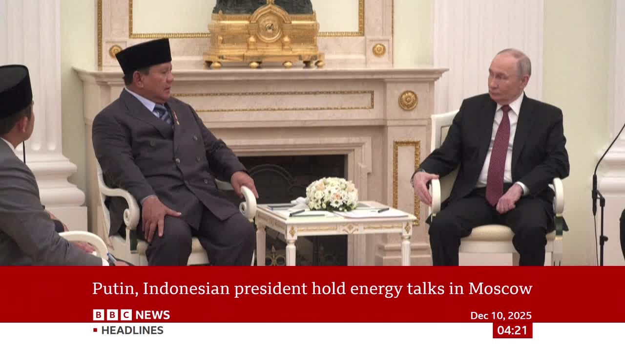 Two men sit in ornate chairs, facing each other across a small table. One man, wearing a dark suit and a red tie, looks intently at the other. The other man, in a dark suit and a peked hat, gestures slightly with his left hand.