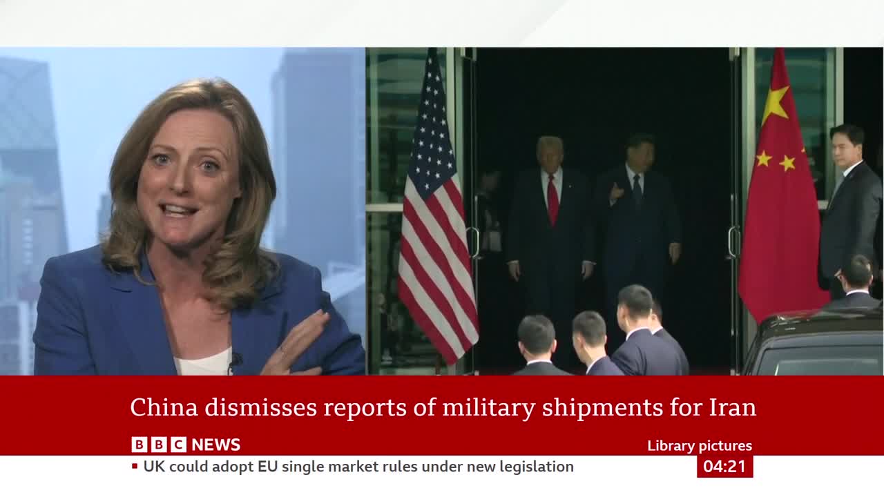 A woman in a blue jacket speaks animatedly on a screen, while another screen shows two men in suits, one with a red tie, walking out of a building. An American flag stands to the left of the doorway, and a Chinese flag is to the right.