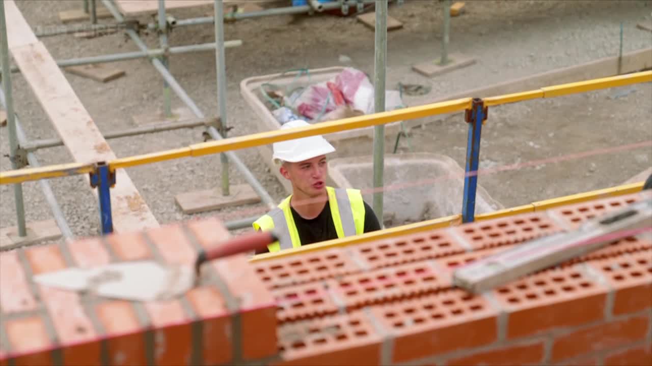 A young man in a hard hat and high-vis vest is looking up from behind a brick wall. A trowel rests on the freshly laid bricks, with a level nearby.
