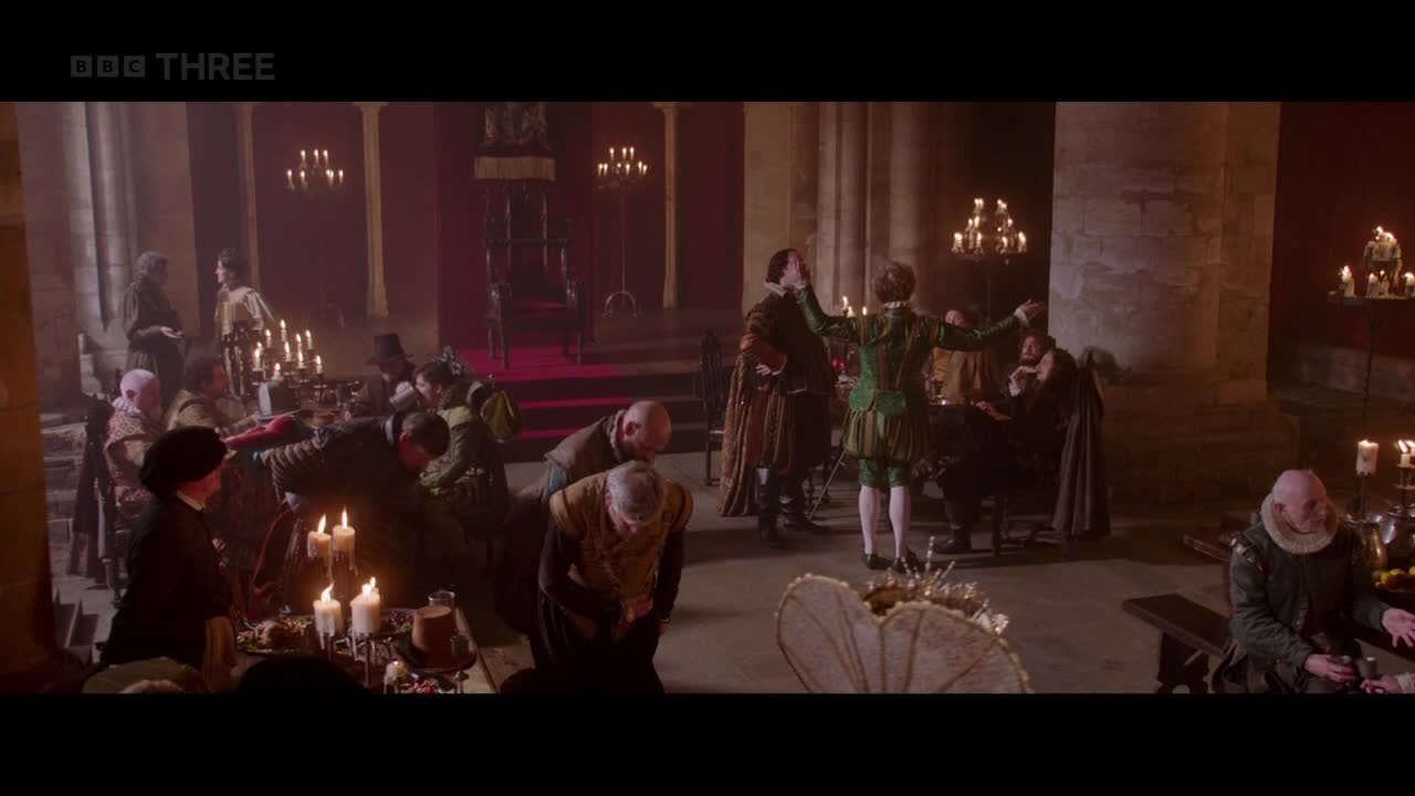 A man in green and gold gestures wildly, arms outstretched, as others watch from candlelit tables. The scene, from BBC Three in the United Kingdom, is set in a grand hall with a raised dais.
