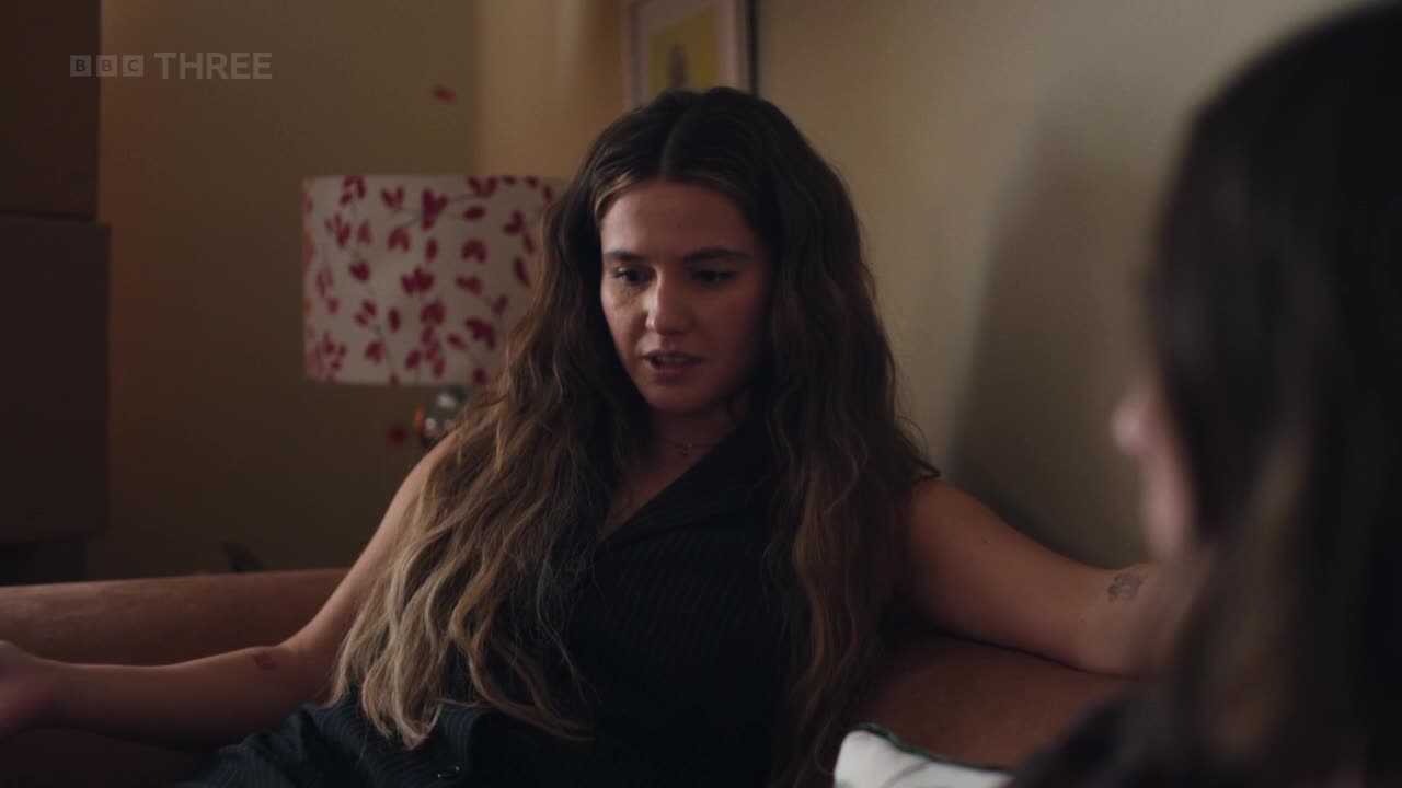 The woman with long, wavy hair is speaking, gesturing with her hands as she sits on the sofa. Her gaze is fixed on someone out of frame, and a BBC Three logo is visible in the corner.
