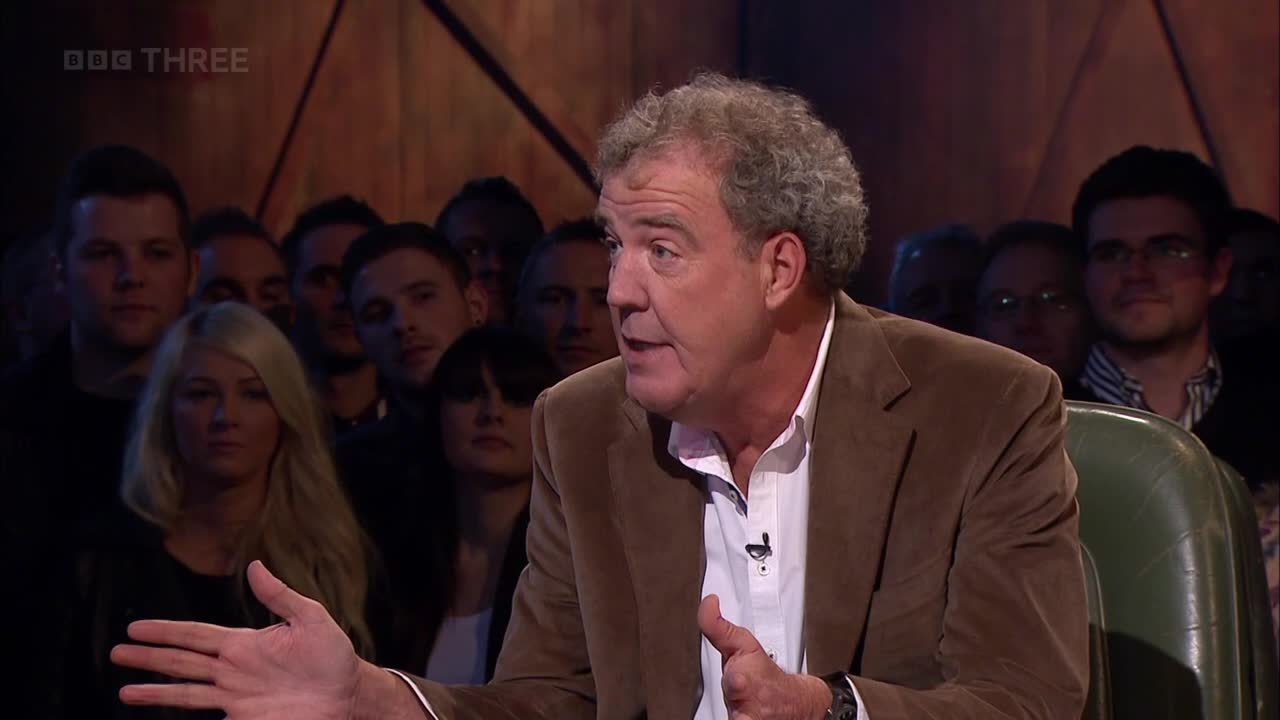 The man in the brown jacket gestures with his left hand, speaking to an unseen audience. Behind him, a crowd of people watches intently, their faces lit by the studio lights.
