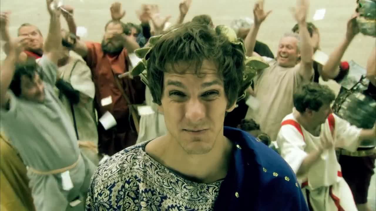 A man in a laurel wreath stares directly at the camera, while a crowd behind him throws confetti and cheers. The scene looks like something from a BBC Three comedy, with the actors in Roman-style robes.
