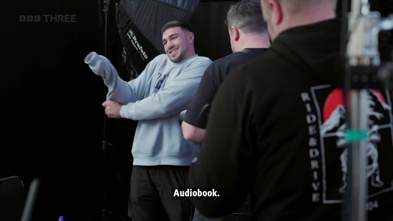 A man in a light blue sweatshirt is pulling up his sleeve, smiling at the camera. Two other men stand nearby, one wearing a black hoodie with a graphic on the back.
