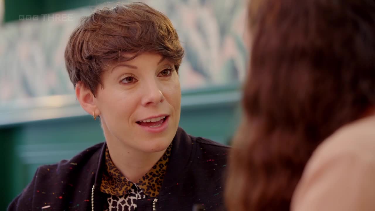 A woman with short brown hair is speaking, her mouth open as if mid-sentence. She's wearing a black jacket over a leopard print shirt, and a small gold hoop earring.
