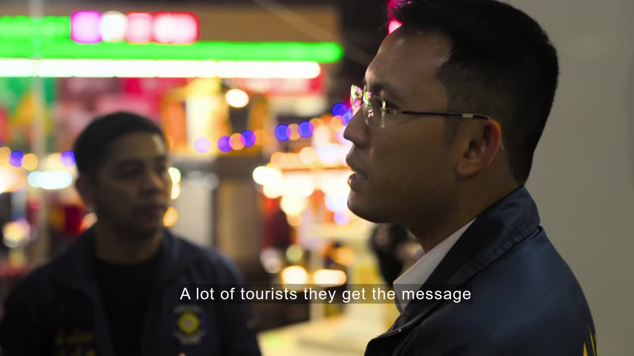 Two men in dark jackets are talking, one wearing glasses and looking off-camera. Behind them, blurred lights from a busy street market create a colorful backdrop.
