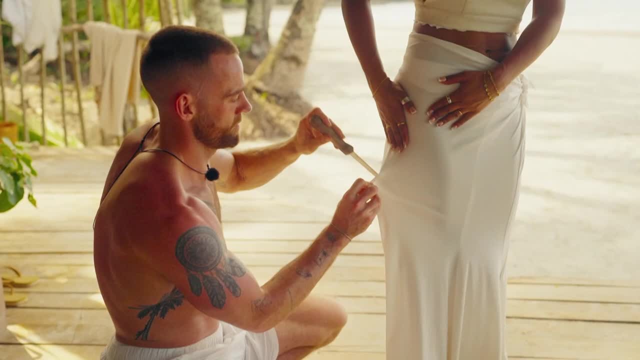 A man, shirtless with tattoos, carefully cuts a piece of fabric on a woman's skirt with a small knife. She stands still, her hands resting on her hips as he works.
