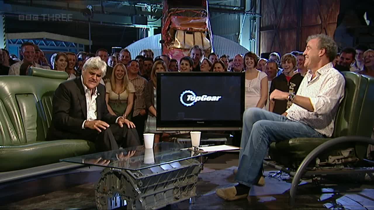 The host in the button-down shirt is chuckling, while the other man, seated next to him, looks on with a slight smile. A crowd of people are gathered behind them, watching the broadcast on BBC Three from the UK.
