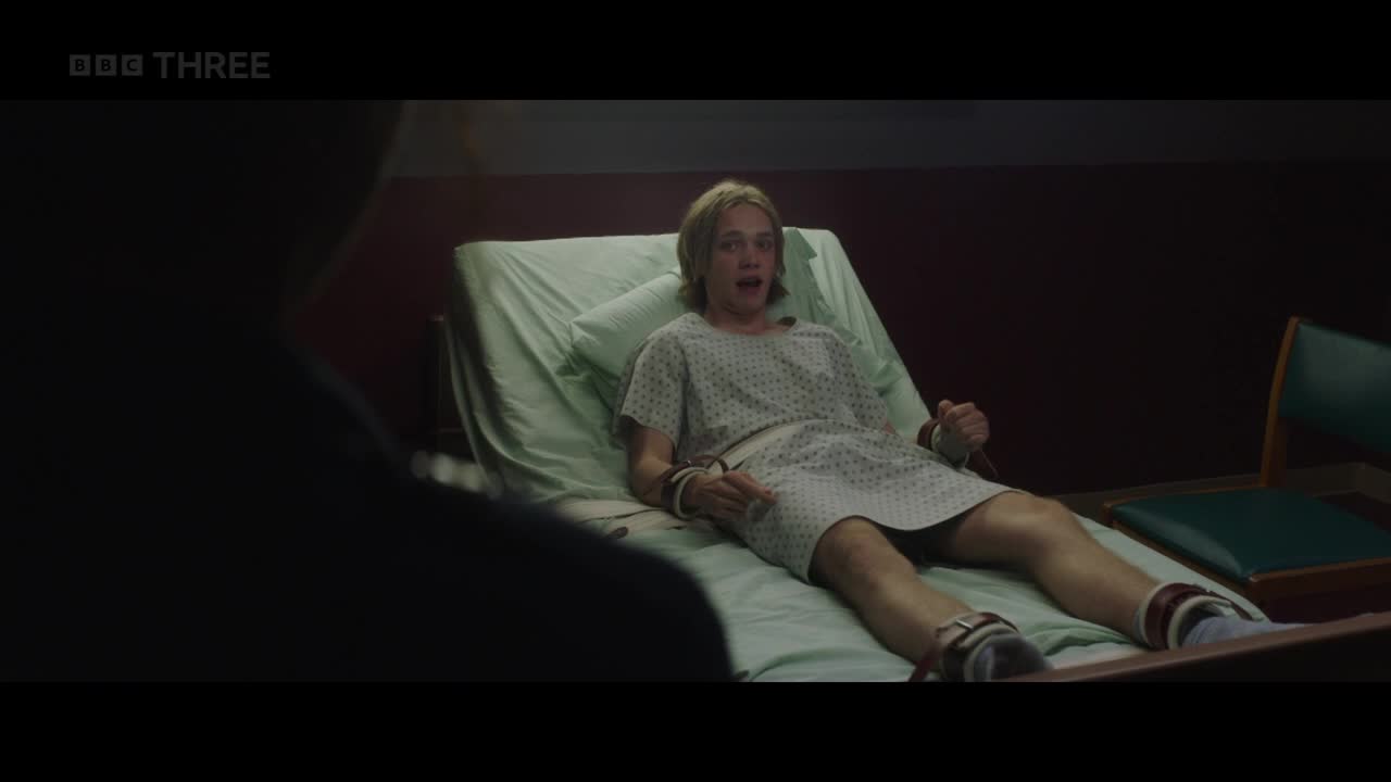 A young person, dressed in a hospital gown, is strapped to a bed, looking startled. Someone is standing in the foreground, their face obscured by shadow.
