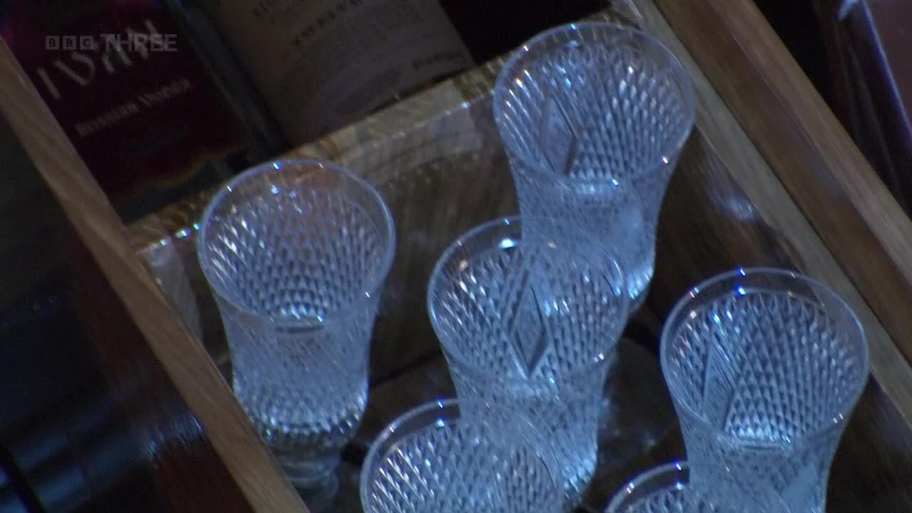 Crystal glasses, etched with a diamond pattern, are stacked in a wooden cabinet. The BBC Three logo is visible in the top corner, hinting at the setting.

