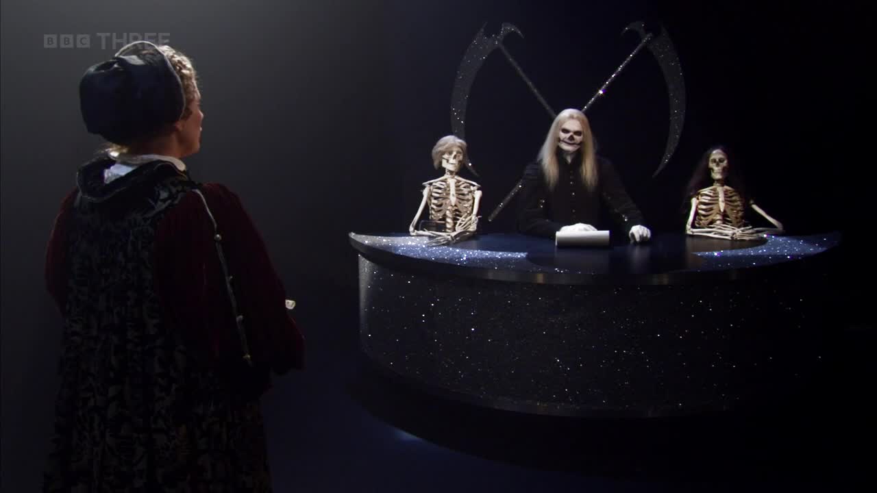 A person in period dress stands facing a desk where three skeletons sit, one with long blonde hair. The BBC Three logo is visible in the corner.
