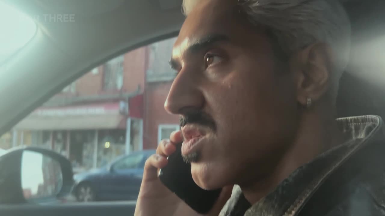 Inside a car, a man with a distinctive mustache and bleached hair is speaking on his phone. Through the window, a street scene in the United Kingdom is visible in the background.
