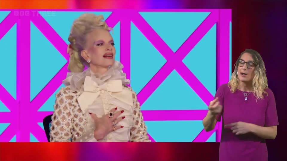 A woman in a dramatic white outfit with a ruffled collar gestures with her hand on her chest. To her right, a woman in a magenta dress signs with her hands.