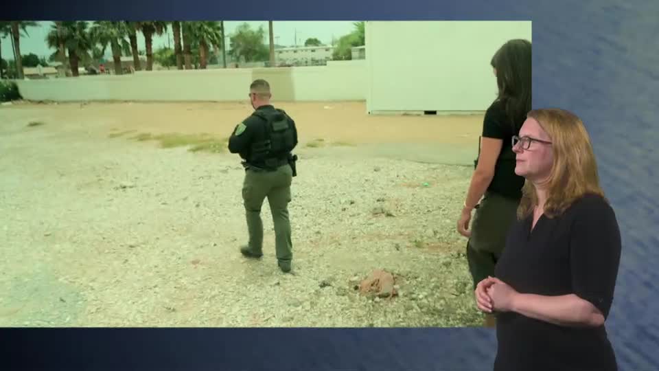 A uniformed officer walks away from the camera across dusty ground. A woman in a black top watches him, her hands clasped.