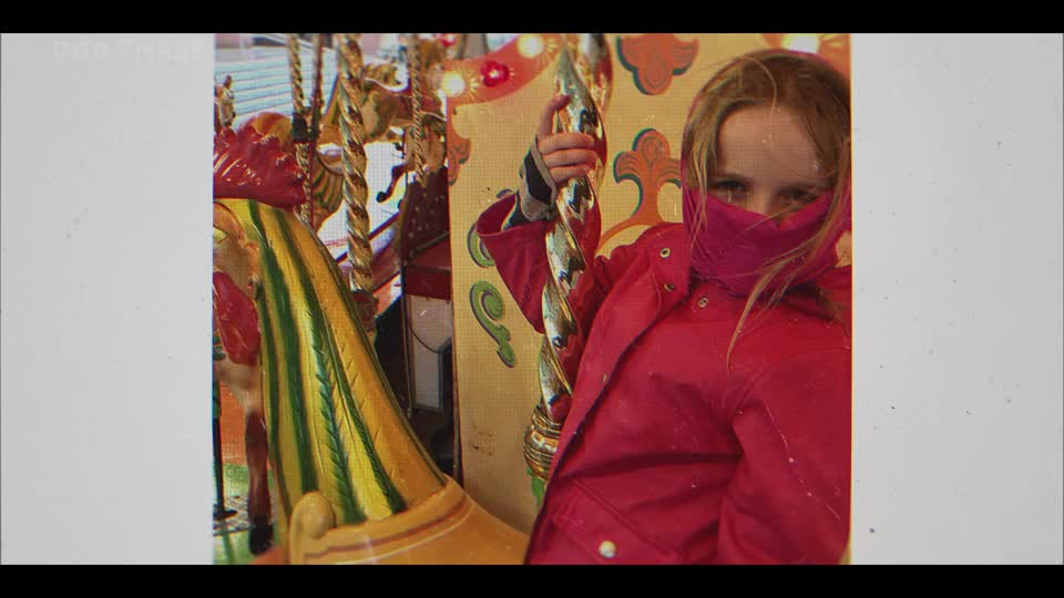 A young girl in a bright red coat grips a golden pole as the carousel spins. Her face is partially obscured by a pink scarf, but her eyes are visible, peeking out at the camera.