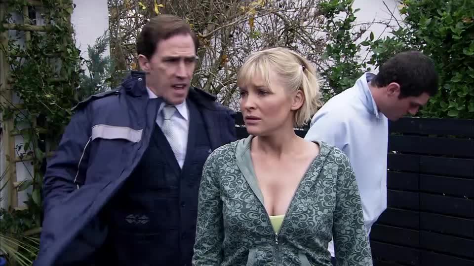 A man in a dark jacket and tie gestures urgently towards a woman in a patterned hoodie. Another man in a light blue sweatshirt bends his head down behind them. This scene is from a BBC Three drama.