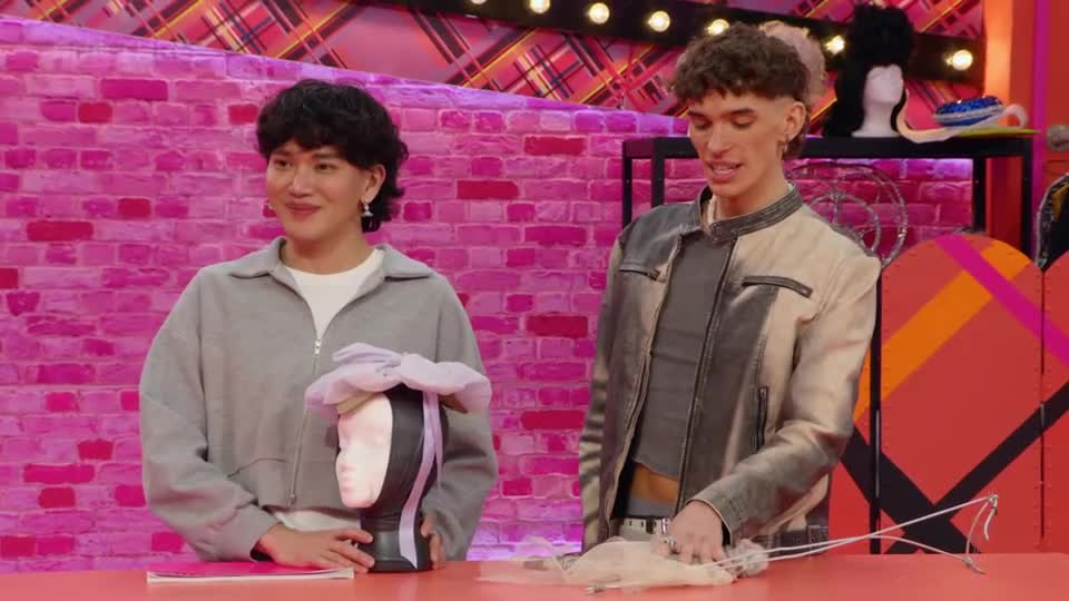 The person on the left smiles, holding a wig stand adorned with a lilac bow. Beside them, the other person gestures towards a delicate, sheer fabric spread across the table.